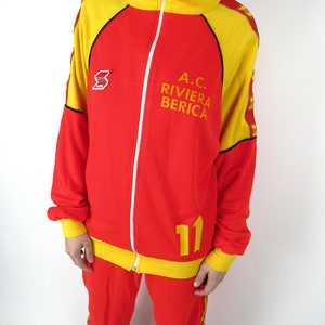 Puede incluir: Ch&aacute;ndal rojo y amarillo con cierre de cremallera. La chaqueta tiene un cuello amarillo y rayas amarillas en las mangas. El pantal&oacute;n tiene rayas amarillas en los laterales. La chaqueta tiene el texto "A.C. RIVIERA BERICA" y el n&uacute;mero "11" en la parte delantera.