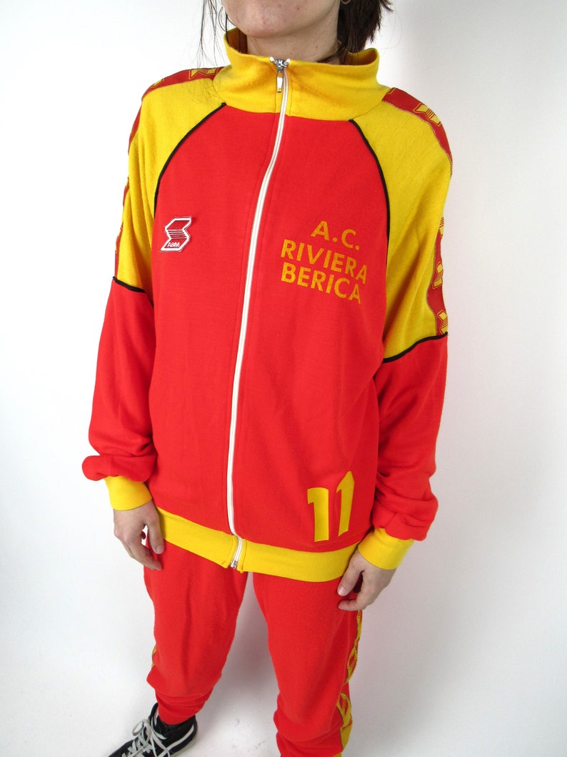 Puede incluir: Ch&aacute;ndal rojo y amarillo con cierre de cremallera. La chaqueta tiene un cuello amarillo y rayas amarillas en las mangas. El pantal&oacute;n tiene rayas amarillas en los laterales. La chaqueta tiene el texto "A.C. RIVIERA BERICA" y el n&uacute;mero "11" en la parte delantera.