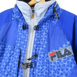 May include: Blue and white FILA ski jacket with a geometric pattern. The jacket has a high collar, a zipper closure, and the FILA logo on the chest. The jacket is hanging on a wooden hanger.