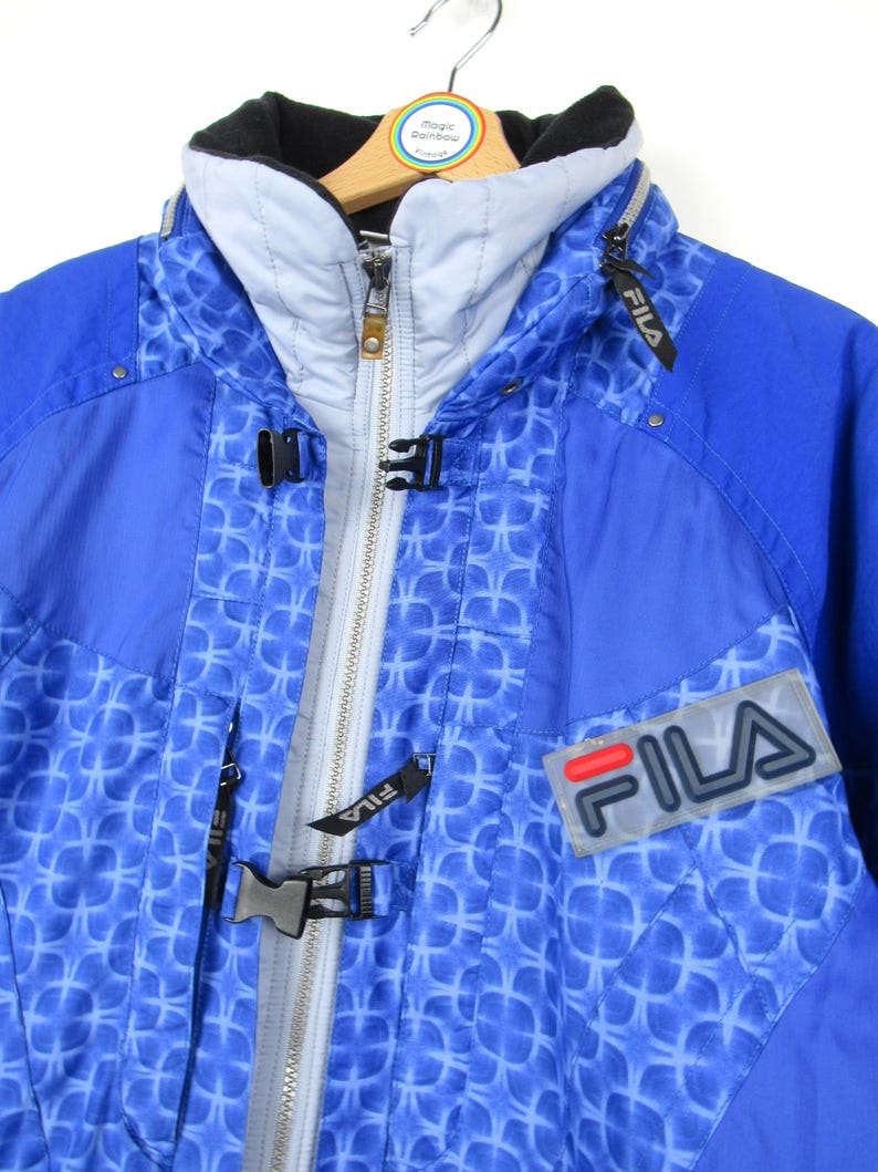 May include: Blue and white FILA ski jacket with a geometric pattern. The jacket has a high collar, a zipper closure, and the FILA logo on the chest. The jacket is hanging on a wooden hanger.