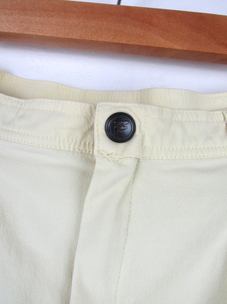 May include: Close-up of a light yellow pair of pants with a black button featuring a stylized letter "F".