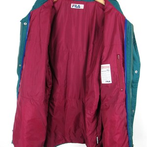May include: A teal blue Fila puffer jacket with a red quilted lining. The jacket has a zipper closure and a snap button closure. The jacket has a Fila logo on the left side of the chest.