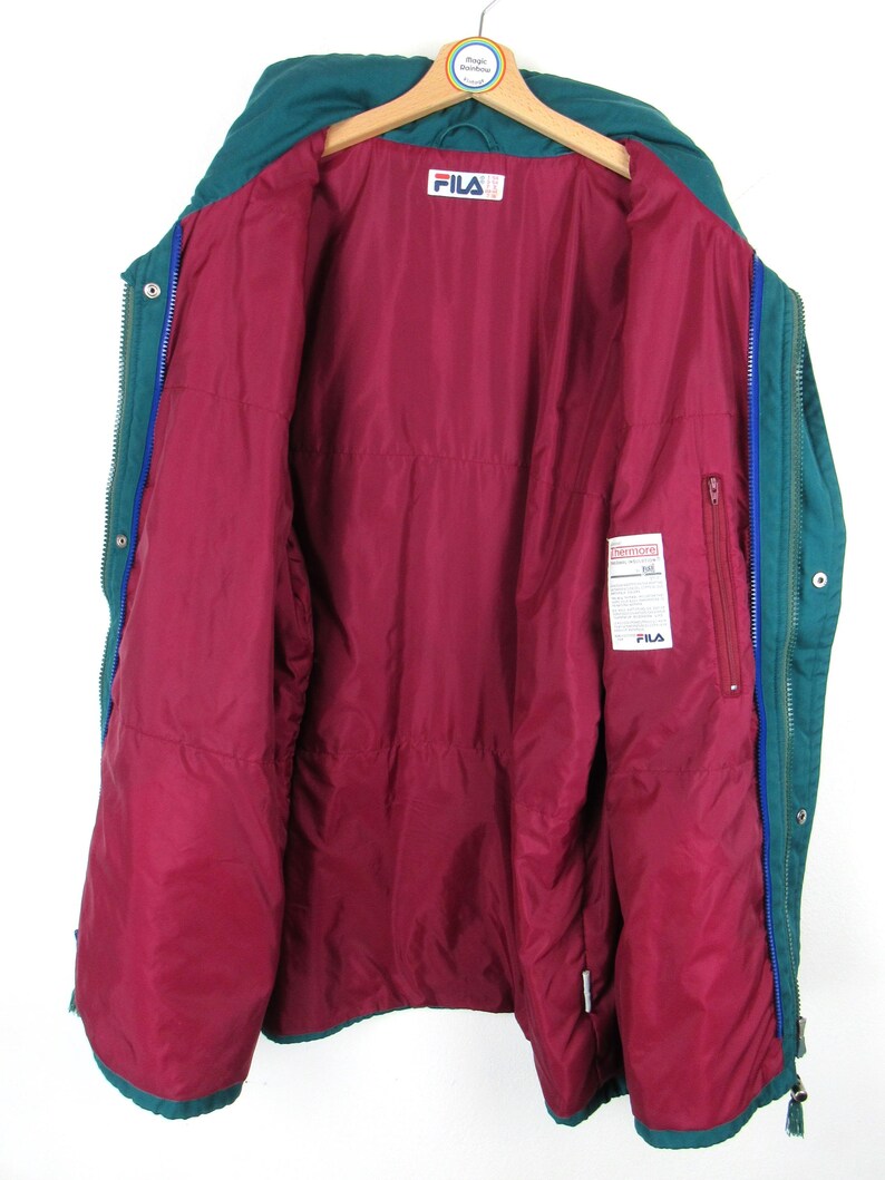 May include: A teal blue Fila puffer jacket with a red quilted lining. The jacket has a zipper closure and a snap button closure. The jacket has a Fila logo on the left side of the chest.