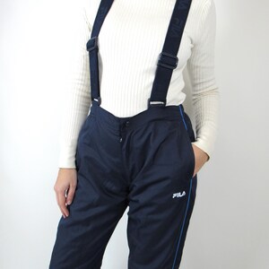May include: Navy blue ski pants with a blue stripe down the side and a white FILA logo. The pants have suspenders with the word FILA repeated on the straps.