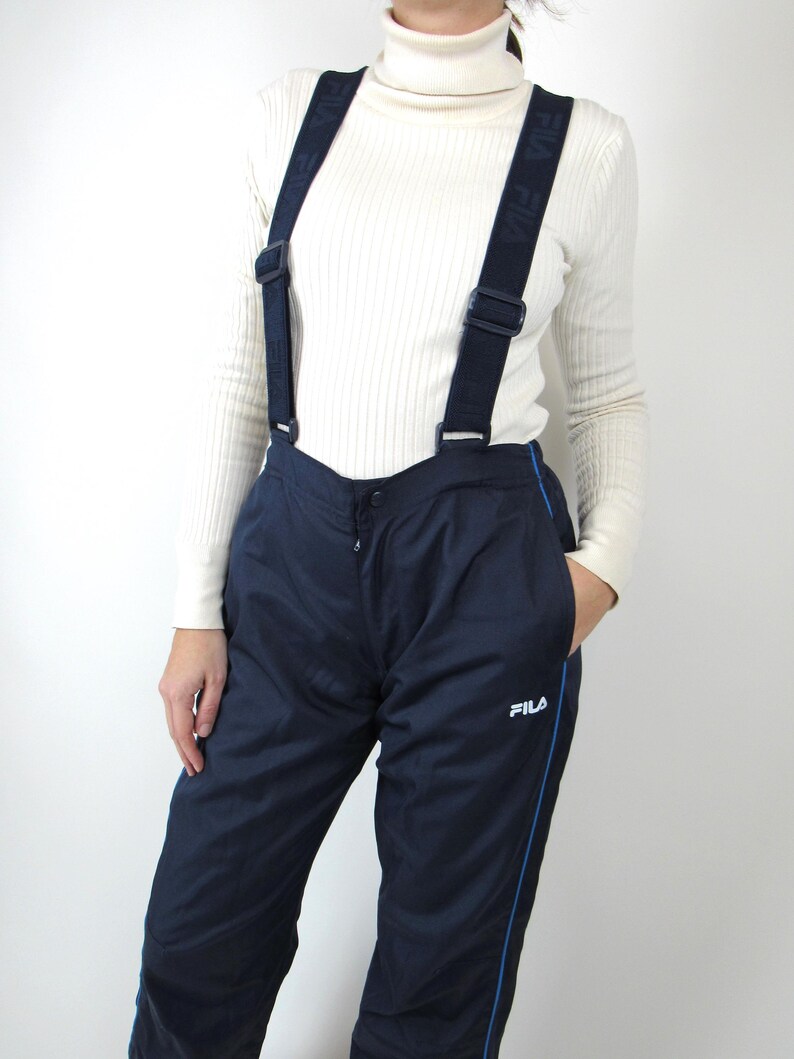 May include: Navy blue ski pants with a blue stripe down the side and a white FILA logo. The pants have suspenders with the word FILA repeated on the straps.