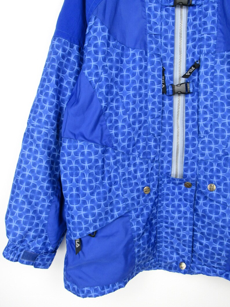 May include: A blue and white patterned jacket with a zipper closure and snap buttons. The jacket has a geometric design and the brand name "FILA" is visible on the zipper and pocket.