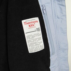 May include: Close-up of a light blue jacket interior with a black lining. A white tag with red text reads "Thermore N.R.G. ENERGY SAVING." The jacket has a zipper and a blue accent at the bottom.