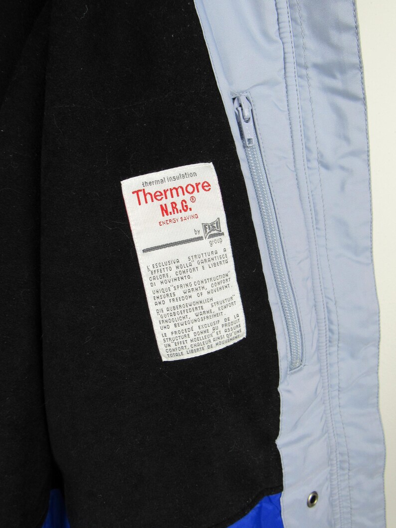 May include: Close-up of a light blue jacket interior with a black lining. A white tag with red text reads "Thermore N.R.G. ENERGY SAVING." The jacket has a zipper and a blue accent at the bottom.