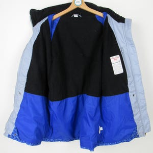 May include: A vintage, open winter jacket with a black upper section, light blue sides, and a vibrant blue lower section. The interior is black, with a tag visible on the right side. The jacket hangs on a wooden hanger against a white background.