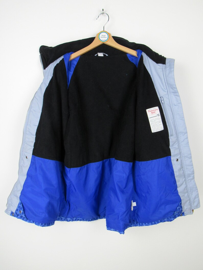 May include: A vintage, open winter jacket with a black upper section, light blue sides, and a vibrant blue lower section. The interior is black, with a tag visible on the right side. The jacket hangs on a wooden hanger against a white background.