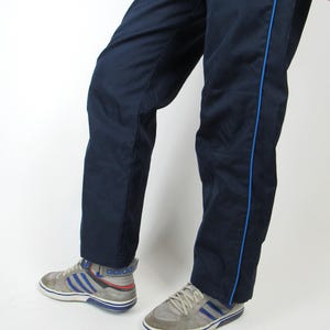 May include: A pair of navy blue sweatpants with blue piping down the sides. The sweatpants are worn by a person wearing gray sneakers with blue accents.