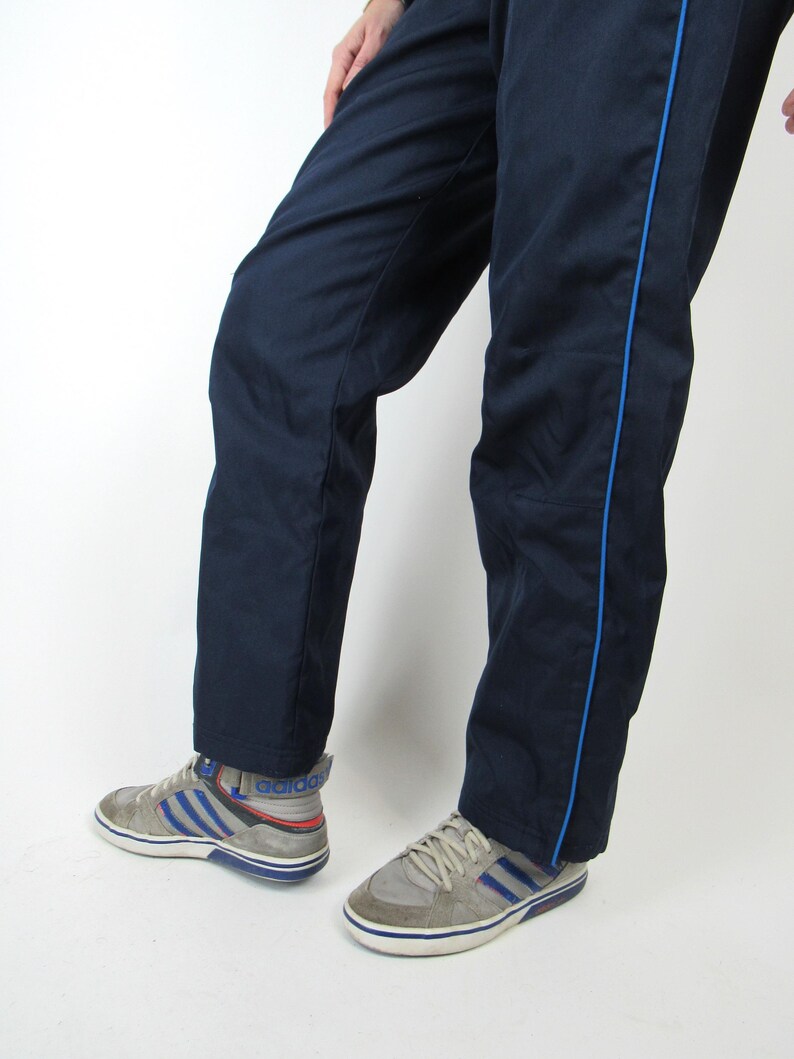 May include: A pair of navy blue sweatpants with blue piping down the sides. The sweatpants are worn by a person wearing gray sneakers with blue accents.