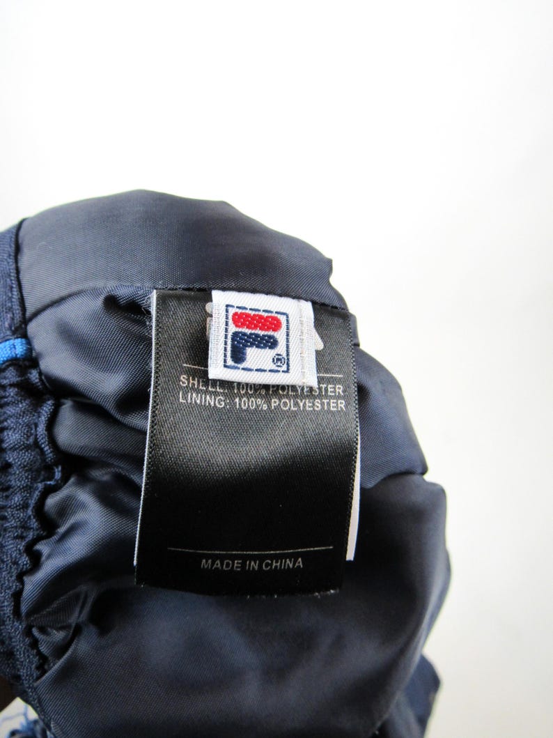 May include: A navy blue and white Fila brand label with the text "SHELL: 100% POLYESTER LINING: 100% POLYESTER MADE IN CHINA".
