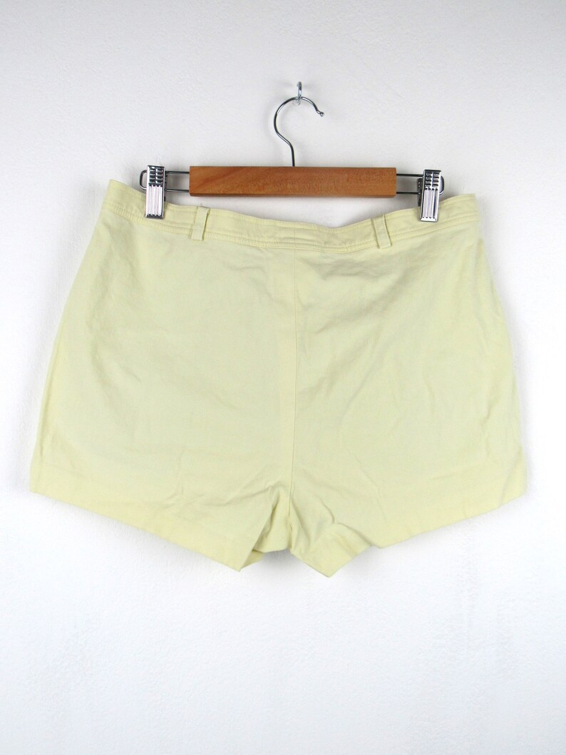May include: A pair of pale yellow shorts hanging on a wooden hanger. The shorts have a simple design and a straight leg.
