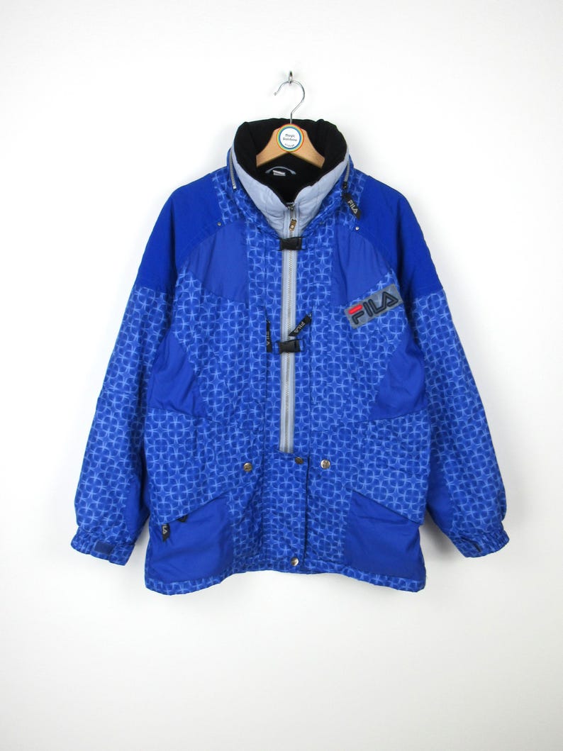 May include: A blue Fila winter jacket with a geometric pattern. The jacket has a black collar, a gray zipper, and the Fila logo on the chest. It features pockets and a hood.