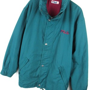 May include: A teal blue Fila jacket with a red lining and a white Fila logo on the front. The jacket has a zipper closure and a button-up flap.