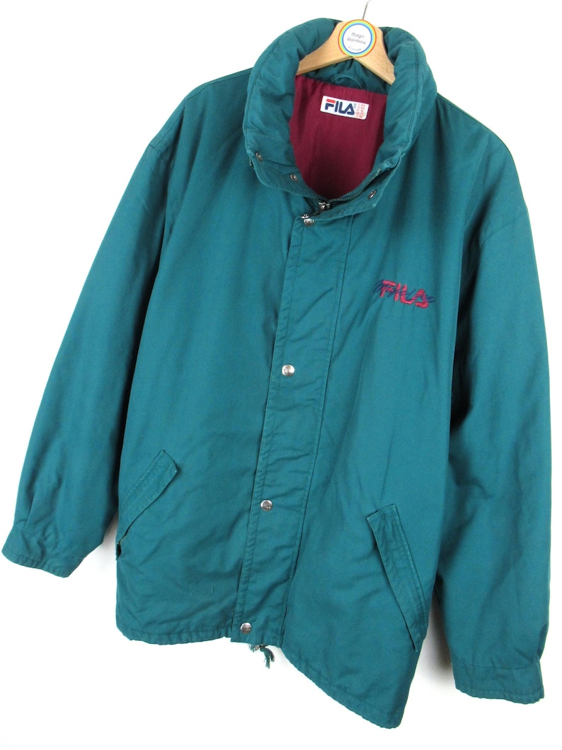May include: A teal blue Fila jacket with a red lining and a white Fila logo on the front. The jacket has a zipper closure and a button-up flap.