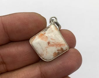 Può includere: Un ciondolo quadrato con una cornice color argento. Il ciondolo presenta una pietra bianca con venature arancioni e marroni. La pietra ha una superficie lucida e un piccolo anello d'argento nella parte superiore per una catena.