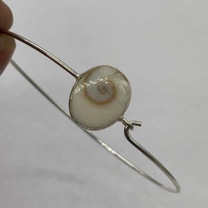 May include: A silver bangle bracelet with a circular, off-white shell pendant displaying a spiral pattern. The pendant is set in a silver frame, and the bracelet has a simple, elegant design. The bracelet is shown against a neutral background.