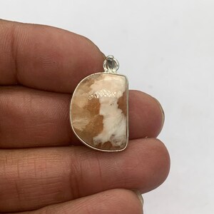 May include: A pendant with a half-moon shape, featuring a silver-colored metal frame. The pendant's interior showcases a mix of beige and white stone, creating a marbled effect. The stone has a polished, glossy finish.