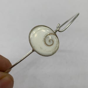 Puede incluir: Una pulsera de brazalete plateada con un colgante de concha blanca en espiral. La concha está engastada en un marco plateado circular. El diseño de la pulsera es sencillo y elegante, adecuado para el uso diario o ocasiones especiales.