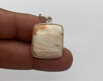 Può includere: Un ciondolo quadrato con una cornice color argento. Il ciondolo presenta una pietra color crema con venature arancioni e bianche. La pietra ha una superficie lucida e un piccolo anello nella parte superiore per una catena. Il ciondolo è tenuto da una mano.