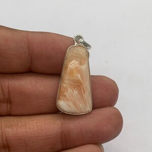 May include: A pendant with a rectangular, trapezoid shape. The pendant features a light orange and white stone set in a silver-colored metal frame. The stone has a unique, textured pattern. The pendant has a small loop at the top for a chain.