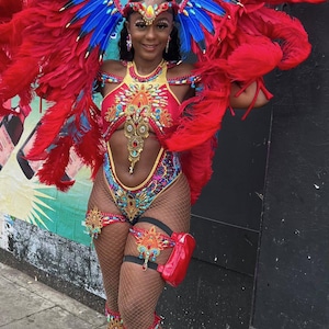 May include: A woman wearing a red and blue feathered headdress, a red and gold sequined costume, and white sandals. She is posing for a photo with a red bag on her thigh.