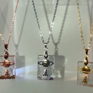 May include: Three necklaces with rectangular pendants featuring a silver rocket and a silver planet with a ring around it. The necklaces are on a white surface. The chains are rose gold, silver, and gold.