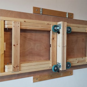 May include: A wooden folding table with wheels attached to the side. The table is folded up against a wall and is secured with a wooden brace.