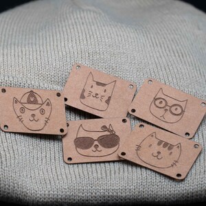 Könnte beinhalten: Fünf braune Leder-Katzen-Patches mit verschiedenen Designs. Jeder Patch hat zwei Löcher an jeder Seite zum Nähen. Die Designs beinhalten eine Katze mit Sonnenbrille, eine Katze mit Baseballkappe, eine Katze mit einem Herz auf dem Kopf, eine Katze mit Brille und eine Katze mit Streifen.