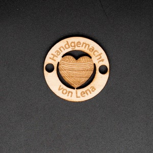 May include: A wooden circle with the text "Handgemacht von Lena" and a heart-shaped cutout filled with a yarn pattern.
