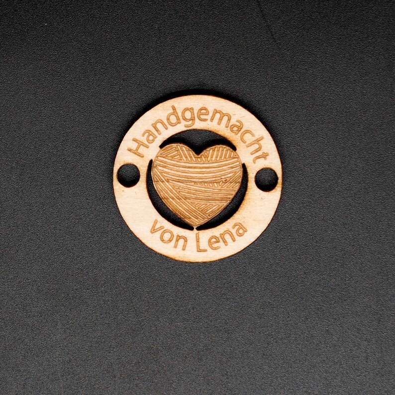 May include: A wooden circle with the text "Handgemacht von Lena" and a heart-shaped cutout filled with a yarn pattern.