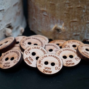 Könnte beinhalten: Nahaufnahme von Holz-Knöpfen mit dem Text "Mit Liebe Handgemacht" und "Von Belle" auf Deutsch. Die Knöpfe sind braun und haben jeweils zwei Löcher. Die Knöpfe liegen auf einem schwarzen Untergrund.