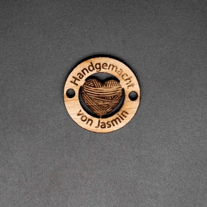 May include: A wooden circle tag with the text "Handgemacht von Jasmin" and a heart-shaped cutout filled with a yarn texture.
