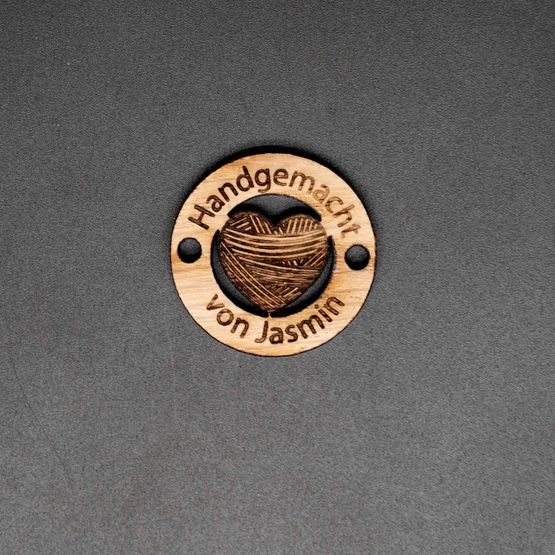 May include: A wooden circle tag with the text "Handgemacht von Jasmin" and a heart-shaped cutout filled with a yarn texture.