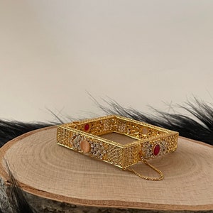 May include: A gold square-shaped bangle with intricate filigree design and clear and red gemstones. The bangle has a chain attached to it.