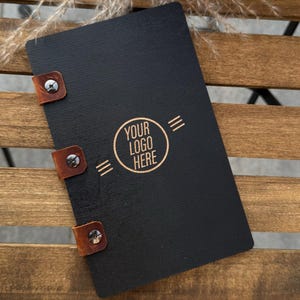 May include: A black menu or notepad with brown leather straps and metal rivets. The cover features the text "YOUR LOGO HERE" in a circular design. The item is displayed on a wooden surface.