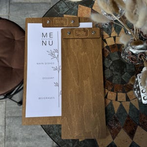 May include: Wooden menu holder with the word "Sicily" at the top. The menu features the words "ME NU" and the categories "MAIN DISHES", "DESSERT", and "BEVERAGES". The holder is made of light brown wood.
