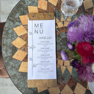Può includere: Un menu della lista dei vini con il testo "Sicily" in alto. Il menu ha una parte superiore e inferiore in legno, con le parole "ME RESTAURANT NU" e "WINE LIST" stampate sopra. Il menu è su un tavolo a mosaico con un bicchiere e dei fiori.