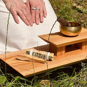 Puede incluir: Una mesa de altar de madera con un cuenco tibetano de latón y un mazo de madera, un manojo de hierbas secas atadas con cuerda blanca y un trozo de madera de palo santo. La mesa tiene una plataforma elevada y un acabado de veta clara.