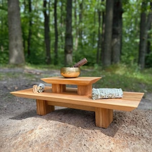 May include: A two-tiered wooden altar stand with a light brown grain finish. A small gold-colored metal singing bowl with a wooden mallet rests on the top tier. A bundle of dried sage and a small piece of wood are placed on the lower tier.