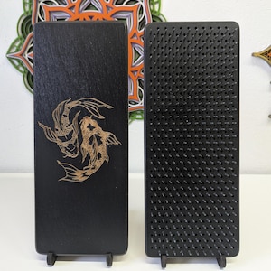 May include: Two black wooden sadhu boards. One board features a gold koi fish design. The other board is covered in black nails. Both boards are rectangular and stand upright on small black supports. A decorative mandala is in the background.