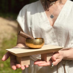 May include: A small, gold-colored metal singing bowl with a hammered texture sits on a light-colored wooden stand with four short legs. A dark brown wooden mallet rests inside the bowl. The bowl features thin horizontal lines etched near the rim.