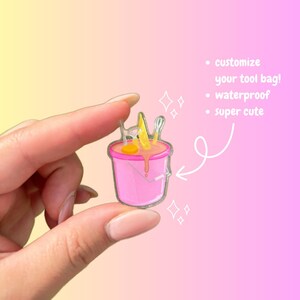 May include: A pink plastic bucket with a yellow lemon, a white whisk, and a pink egg yolk inside. The bucket is being held by a hand. The text "customize your tool bag! waterproof super cute" is written in white on a pink background.