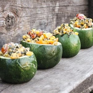 May include: Four green zucchini halves filled with a mixture of corn, spinach, cheese, and other ingredients. The zucchini halves are arranged on a wooden surface.