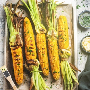 May include: Five ears of grilled corn on the cob, with husks partially removed, on a baking sheet. The corn is yellow and has grill marks. There is parsley scattered on the baking sheet.