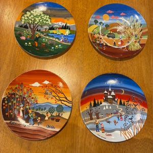 May include: Four round decorative plates featuring colorful seasonal illustrations. The top left depicts spring with a blooming tree, the top right shows a summer harvest, the bottom left displays an autumn grape harvest, and the bottom right shows a winter scene with a castle and people ice skating.