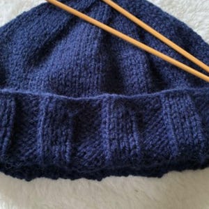 Peut inclure: Un bonnet tricoté bleu foncé avec un revers côtelé et deux aiguilles à tricoter en bois posées dessus. Le texte Fisherman's Beanie est imprimé en haut et le texte Knit it in an evening! est imprimé en bas de l'image.