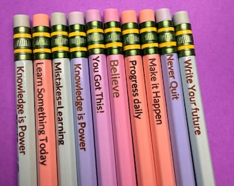 May include: A collection of ten pencils in various pastel colors, each with a motivational phrase printed on the side. The pencils have yellow and green accents near the eraser and lead. The background is a solid purple.